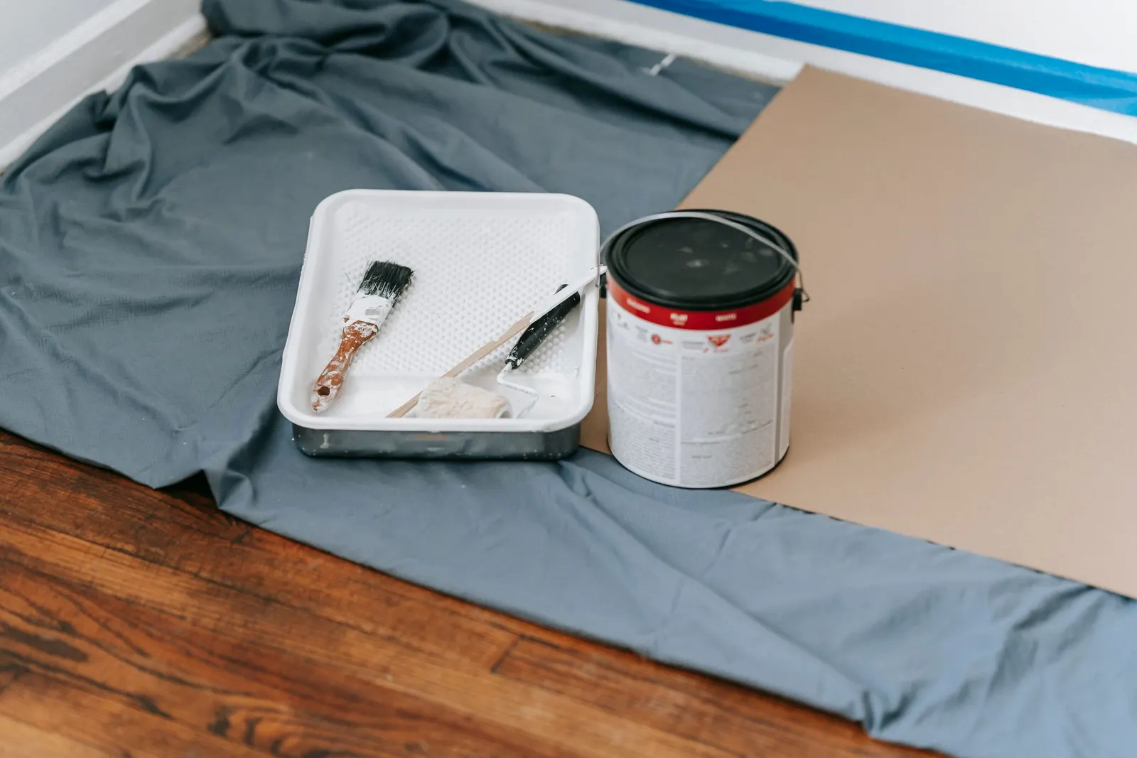 A paint can, tray and brush resting on a drop cloth over a hardwood floor.