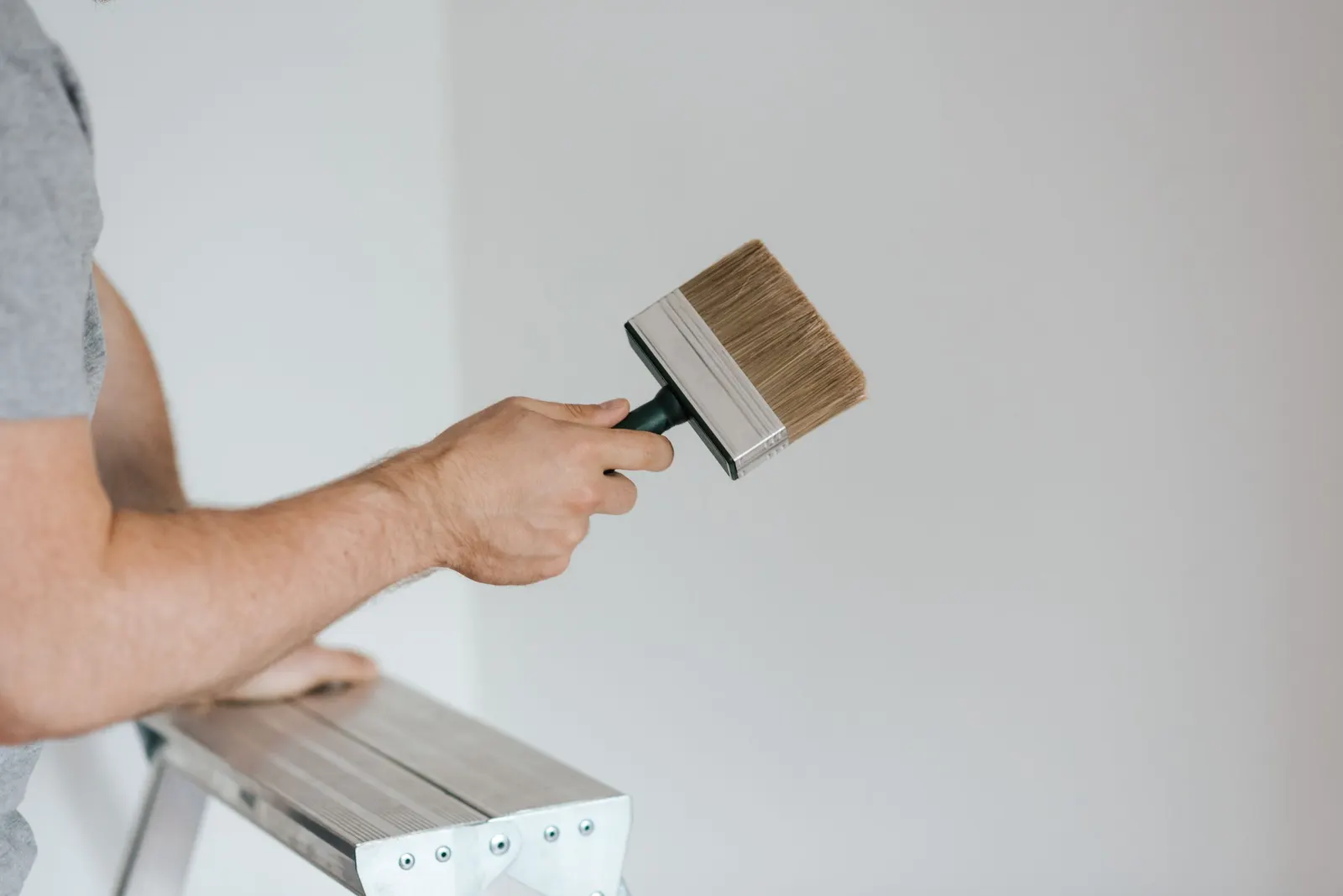 A painter holding a wide brush against a white interior wall beside a step ladder.