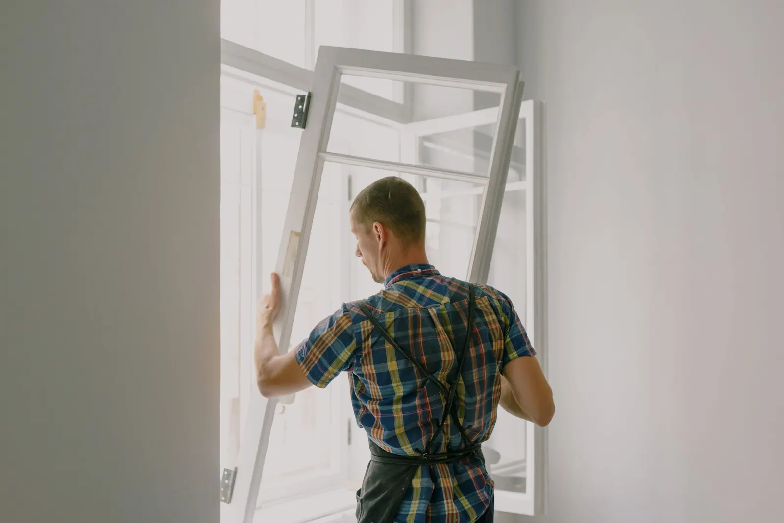 A worker carrying a new white window frame into a room during a window replacement.